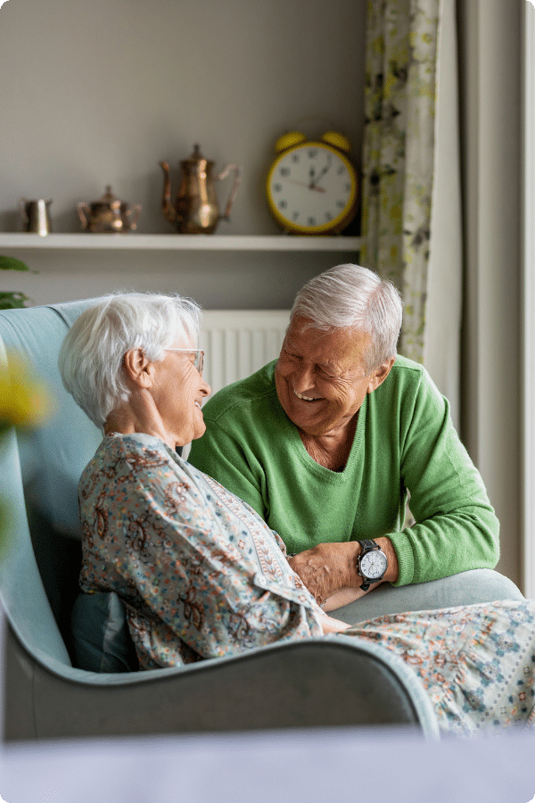 An elderly couple sits together in a living room, smiling and holding hands. The man is wearing a green sweater and the woman a floral dress. A clock and teapots are visible on a shelf in the background.