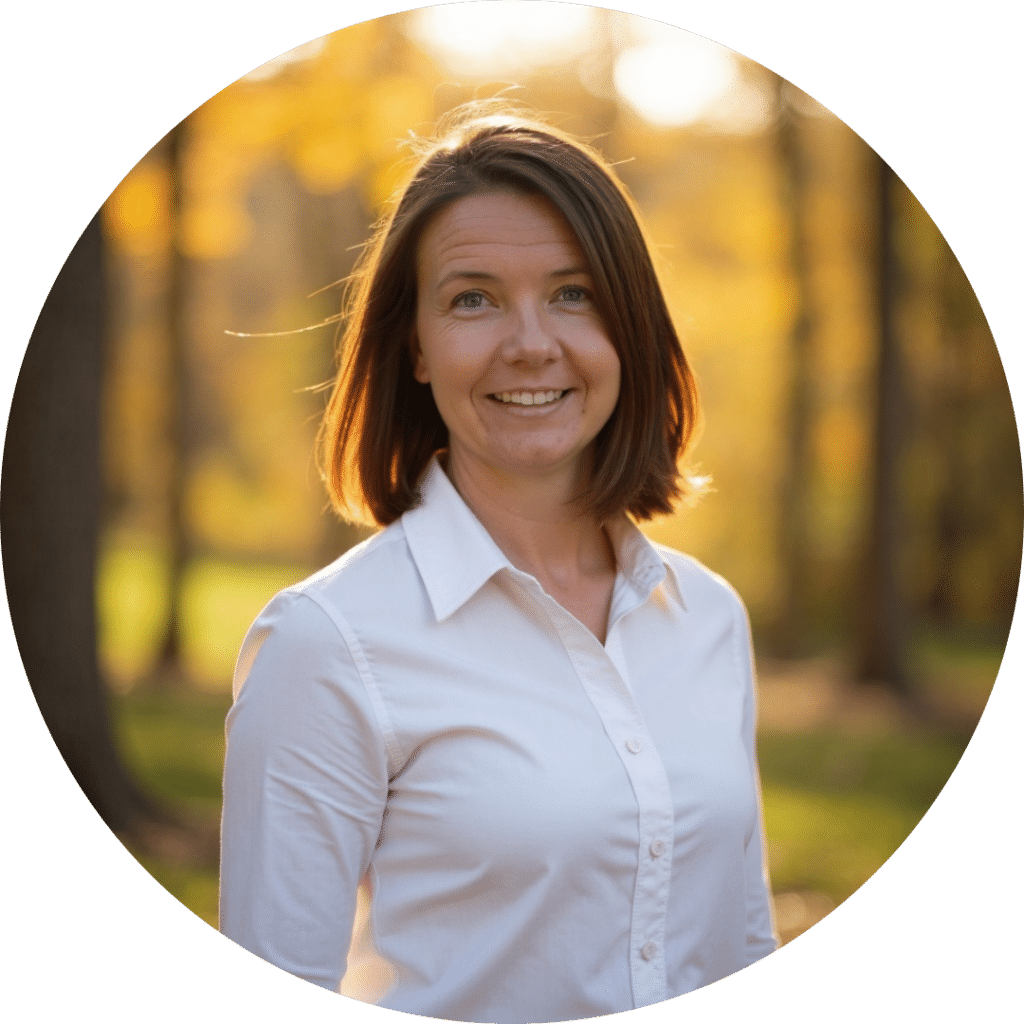 A woman with straight brown hair and a white shirt stands outdoors in a sunlit wooded area, smiling at the camera—radiating warmth much like quality care home health brings to loved ones.