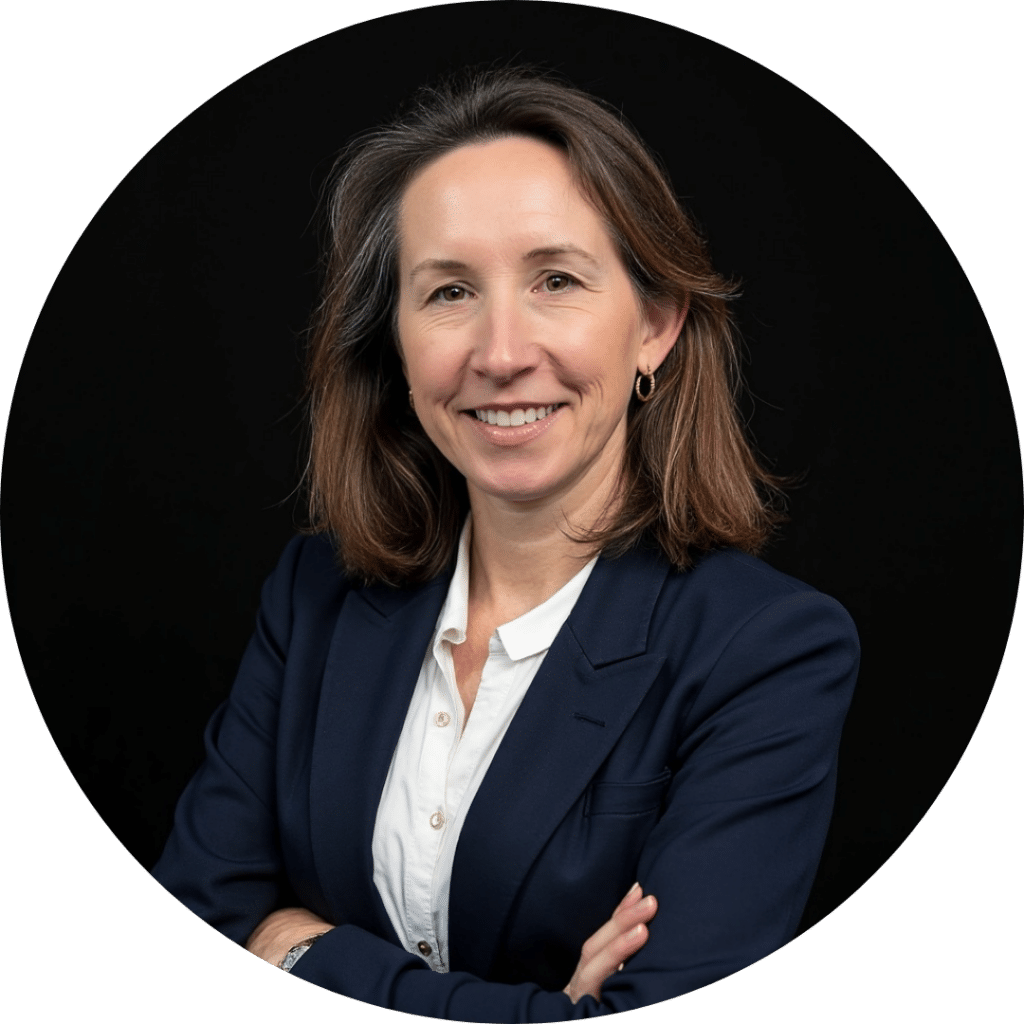 A woman with shoulder-length brown hair, wearing a navy blazer and white shirt, stands with arms crossed against a black background, embodying the confidence and professionalism of quality care home health services.